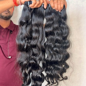 May include: Two bundles of long, wavy, jet-black hair extensions. The hair has a natural, glossy sheen and a consistent, loose wave pattern throughout the length of the strands. Each bundle is secured at the top with a reinforced weft band.
