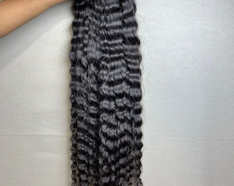 Könnte beinhalten: Ein Bündel dunkler, gewellter Haarverlängerungen. Das Haar hat eine tiefschwarze Farbe mit einem natürlichen Wellenmuster. Die Extensions werden oben zusammengehalten, wobei die welligen Strähnen nach unten fallen. Der Hintergrund ist ein schlichtes, hellgraues.
