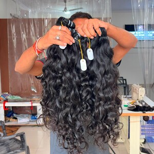 May include: A bundle of long, wavy, black hair extensions held up by the top weft. The hair features a deep, dark colour with a natural-looking wave pattern throughout the length. Several small white tags are attached to the top of the hair bundle.