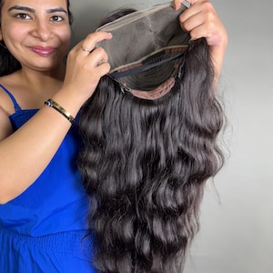 Może przedstawiać: Czarna peruka z włosów ludzkich z falowanymi włosami, pokazana na czapce z koronki. Perukę trzyma osoba w niebieskim kombinezonie.