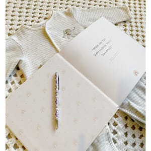 May include: A white baby onesie with gray stripes and an embroidered elephant design is laid out on a white crocheted blanket. A white journal with the text "These are the days I never want to forget" is open on the blanket. A purple and silver pen is lying on the journal's open page, which is decorated with a repeating pattern of rainbow arches.