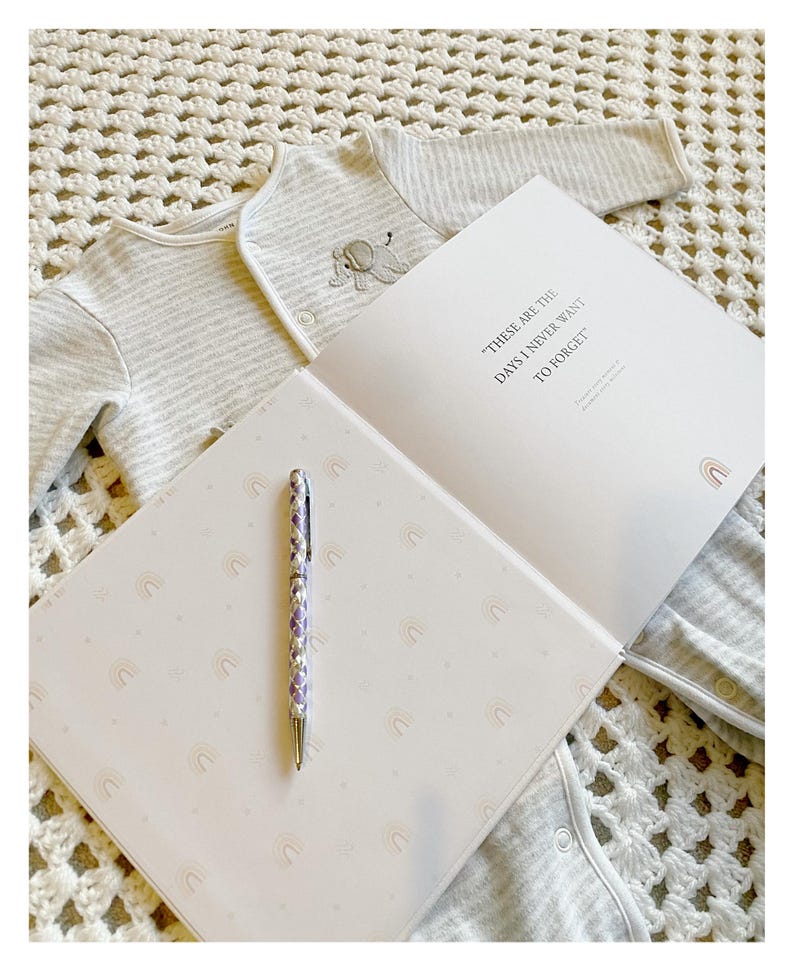 May include: A white baby onesie with gray stripes and an embroidered elephant design is laid out on a white crocheted blanket. A white journal with the text "These are the days I never want to forget" is open on the blanket. A purple and silver pen is lying on the journal's open page, which is decorated with a repeating pattern of rainbow arches.