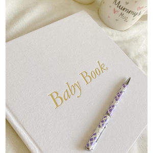 May include: A white baby book with gold lettering that reads "Baby Book". A purple and white pen is lying on top of the book.