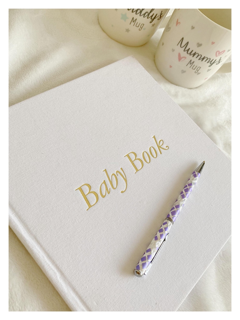 May include: A white baby book with gold lettering that reads "Baby Book". A purple and white pen is lying on top of the book.