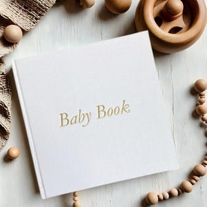 May include: A white baby book with the words "Baby Book" in gold lettering on the cover. The book is surrounded by wooden toys and beads.