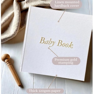 May include: A white linen-covered hardcover baby book with gold foil lettering that reads "Baby Book". The book is on a wooden surface with a wooden dowel next to it. The book has thick 120gsm paper.
