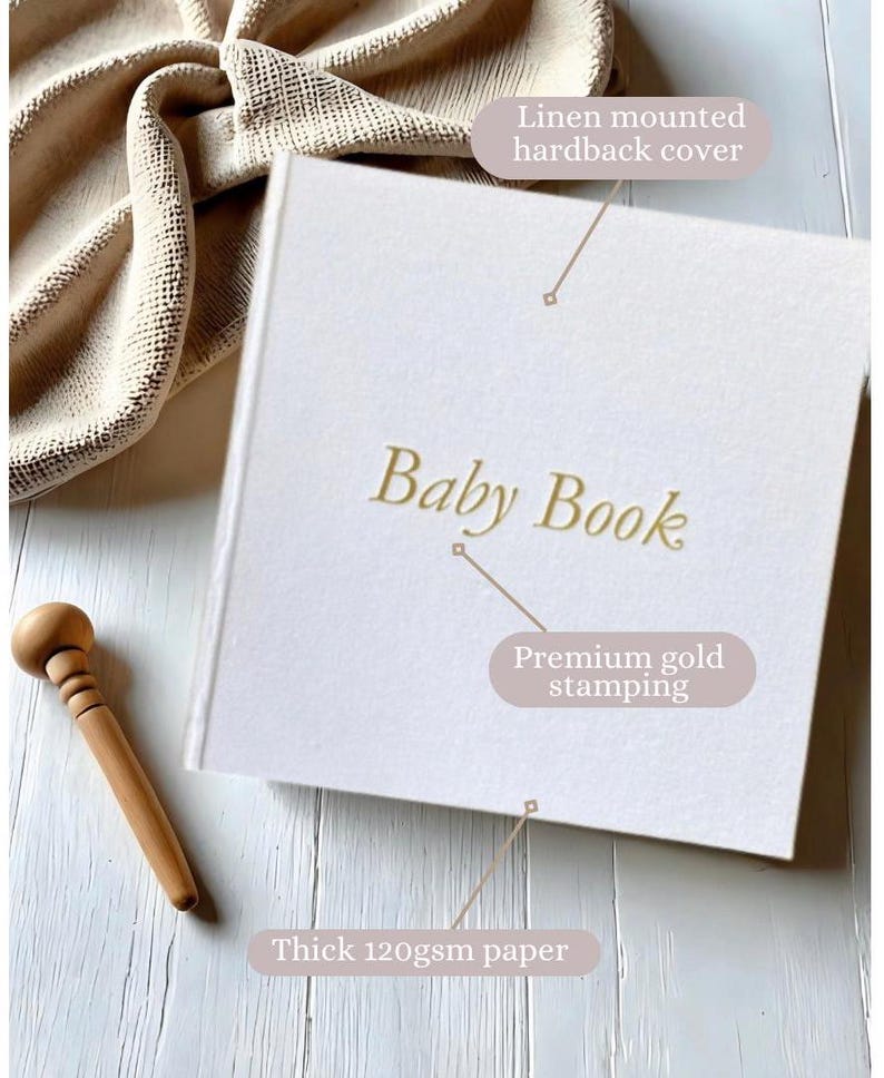 May include: A white linen-covered hardcover baby book with gold foil lettering that reads "Baby Book". The book is on a wooden surface with a wooden dowel next to it. The book has thick 120gsm paper.