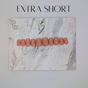 May include: A set of extra short, peach-colored press-on nails arranged on a marble-patterned surface. The nails are neatly aligned, showcasing a uniform shape and color. The text "EXTRA SHORT" is displayed above the nails, with the "Barre V" logo.