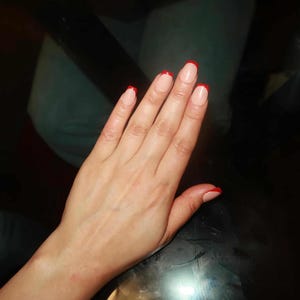 May include: A hand with a French manicure. The nails are painted with a natural base and a bright red tip. The hand is resting on a dark, reflective surface.