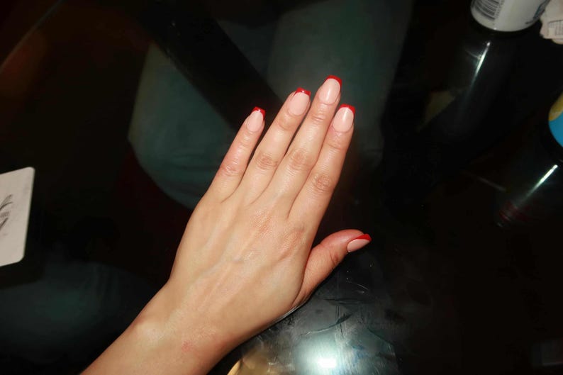 May include: A hand with a French manicure. The nails are painted with a natural base and a bright red tip. The hand is resting on a dark, reflective surface.