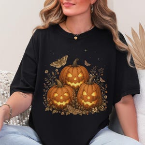 May include: A black short-sleeved t-shirt featuring a graphic print of three glowing jack-o'-lanterns surrounded by small flowers, leaves, and moths. The pumpkins are orange with carved faces, and the design includes small star accents above the pumpkins.
