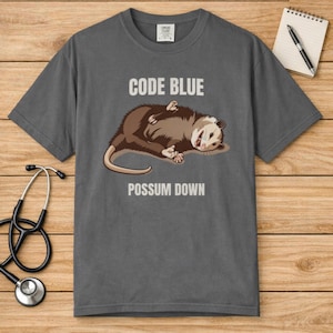 May include: A gray t-shirt with the words "CODE BLUE" and "POSSUM DOWN" printed on it. The shirt features a cartoon opossum lying on its back. A stethoscope and notepad with a pen are on the wooden surface.