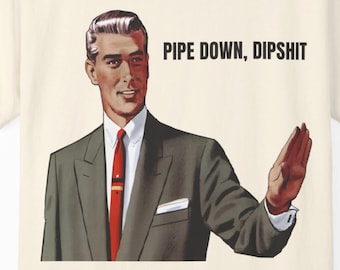 May include: Cream-colored t-shirt featuring a vintage illustration of a man in a suit, red tie, and white shirt. The text "PIPE DOWN, DIPSHIT" is printed above the man's head in black capital letters. The man is gesturing with his hand.