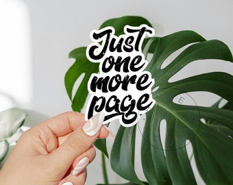 Può includere: Un adesivo bianco con la scritta "Just one more page" in nero, con un carattere invecchiato. L'adesivo è tenuto davanti a una grande foglia verde, con uno sfondo bianco.