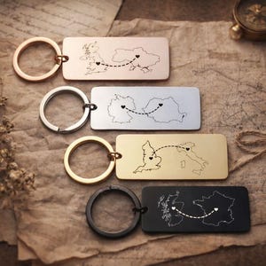 May include: Four rectangular keyrings with engraved map outlines and heart markers connected by dotted lines. The keyrings are in gold, silver, and black, with a brass key ring. The maps depict different countries and routes.