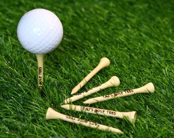 Può includere: Cinque tee da golf in legno con la scritta "Tan's Golf Tees" stampata su di essi. Un tee tiene una palla da golf bianca.
