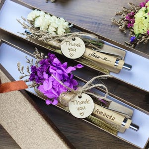 Peut inclure: Deux stylos en bois, chacun dans une boîte cadeau, sont ornés de fleurs séchées et d'une petite étiquette en bois avec "Merci" et un nom. Un stylo a des fleurs violettes, l'autre des fleurs blanches. Les boîtes sont dorées.