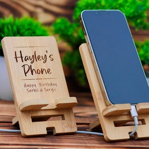 May include: Two wooden phone stands. One stand has the text "Hayley's Phone" and "Happy Birthday Serhii & Sergii" engraved on it. The other stand has a black smartphone in it.