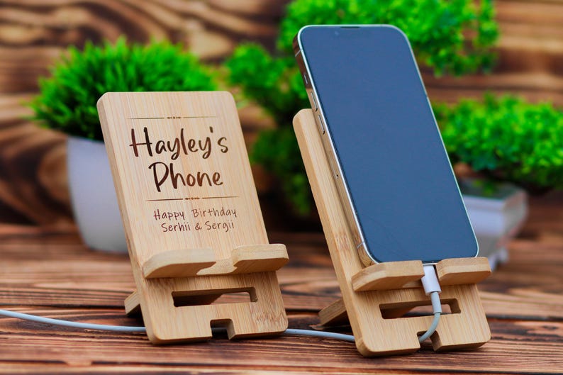 May include: Two wooden phone stands. One stand has the text "Hayley's Phone" and "Happy Birthday Serhii & Sergii" engraved on it. The other stand has a black smartphone in it.