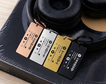 May include: Four rectangular metal keychains in rose gold, silver, gold, and black finishes. Each keychain features a laser-engraved music player interface with a play button, track progress bar, and a scannable sound wave code graphic.
