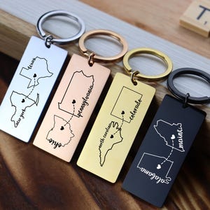 Könnte beinhalten: Schlüsselanhänger mit rechteckigen Metallanhängern in Silber, Roségold, Gold und Schwarz. Jeder Schlüsselanhänger zeigt eine Umrisslinie eines Bundesstaates mit einer gepunkteten Linie und einem Herz sowie einen Staatsnamen. Die Schlüsselanhänger liegen auf einer Holzoberfläche.