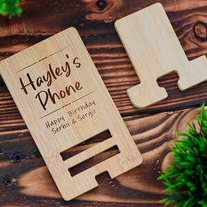 May include: A personalized bamboo phone stand with the text "Hayley's Phone" and "Happy Birthday Serhii & Sergii" engraved on it.