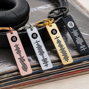 Op de afbeelding: Vier rechthoekige metalen sleutelhangers in roségoud, zilver, goud en zwarte afwerkingen. Elke sleutelhanger heeft een cirkelvormig Spotify-logo bovenaan en een verticaal gegraveerde geluidsgolfgrafiek, bevestigd aan een bijpassende metalen ring.