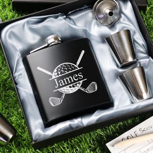 Op de afbeelding: Een zwarte heupfles met een golfbal- en clubontwerp, gepersonaliseerd met de naam "James", wordt gepresenteerd in een geschenkdoos met twee shotglaasjes, een trechter en twee golfballen. De set wordt gepresenteerd op een satijnen bed.
