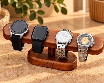 Op de afbeelding: Een houten horlogestandaard met een horizontale balk en een voet met de gegraveerde initialen J.B. Vier polshorloges worden op de balk getoond: een zwarte smartwatch met een gestructureerde band, een zwarte vierkante smartwatch en twee zilveren horloges.