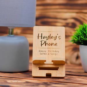 May include: A wooden phone stand with the text "Hayley's Phone" and "Happy Birthday Serhii & Sergi!" engraved on it. The stand is made of light brown wood and has a slot for holding a phone.