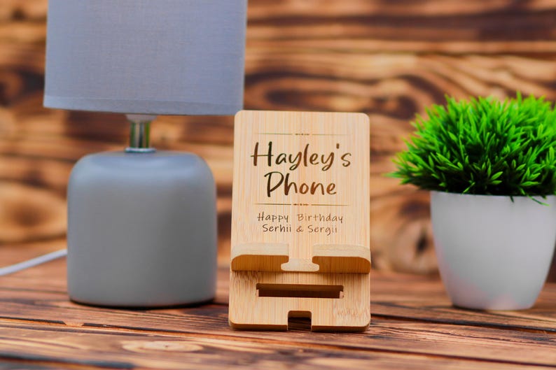May include: A wooden phone stand with the text "Hayley's Phone" and "Happy Birthday Serhii & Sergi!" engraved on it. The stand is made of light brown wood and has a slot for holding a phone.