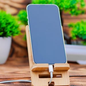 May include: A black smartphone with a white charging cable plugged into the bottom. The phone is in a wooden stand on a wooden table.