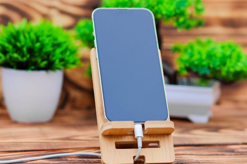 May include: A black smartphone with a white charging cable plugged into the bottom. The phone is in a wooden stand on a wooden table.