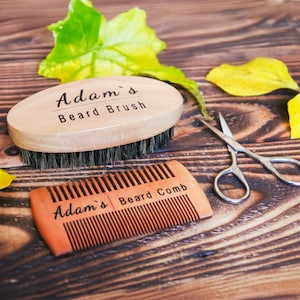 May include: A wooden beard brush and comb set with the text "Adam's" and "Beard Brush" and "Beard Comb" printed on them. A pair of small silver scissors and some yellow and green leaves are also in the image. The items are on a wooden surface.