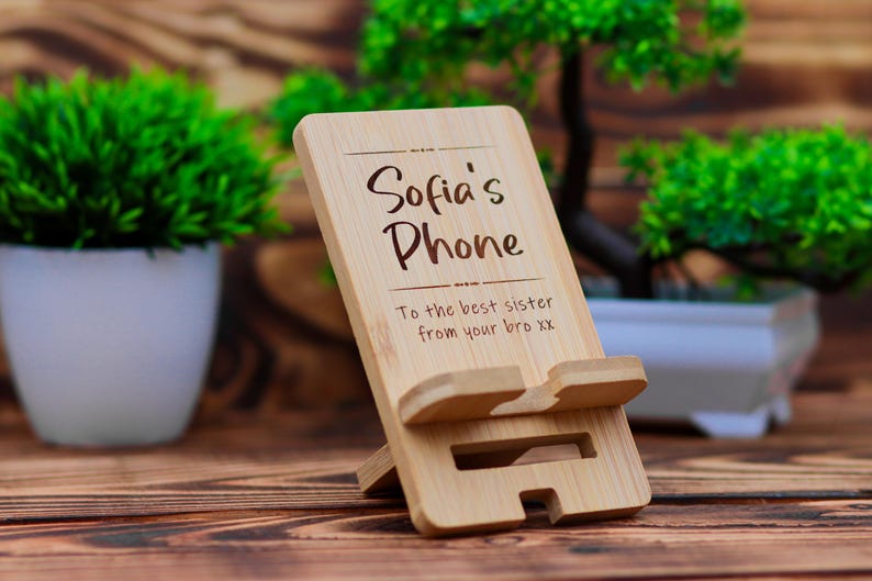 May include: A wooden phone stand with the text "Sofia's Phone" and "To the best sister from your bro xx" engraved on it. The stand is made of natural bamboo and has a simple design.