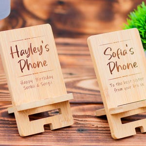 May include: Two wooden phone stands with personalized messages. The first stand says "Hayley's Phone Happy Birthday Serhii & Sergi". The second stand says "Sofia's Phone To the best sister from your bro xx".