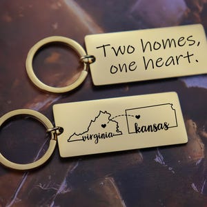 Könnte beinhalten: Zwei goldene, rechteckige Schlüsselanhänger. Einer hat den Text "Two homes, one heart". Der andere zeigt eine Karte von Virginia und Kansas mit Herzen und den Namen der Bundesstaaten.