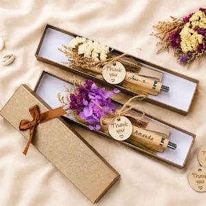 Könnte beinhalten: Zwei rechteckige braune Geschenkboxen mit Holzstiften, die mit Trockenblumen verziert sind. Jeder Stift hat ein rundes Holzschild mit dem Text Thank you und einem Herzsymbol, wobei die Namen Jess und Amanda in den Stift eingraviert sind.