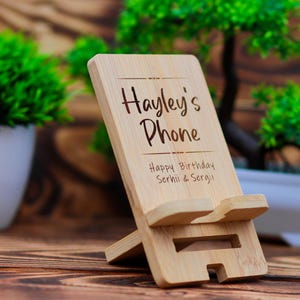 May include: A wooden phone stand with the text "Hayley's Phone" and "Happy Birthday Serhii & Sergii" engraved on it. The stand is made of natural bamboo and has a simple design.