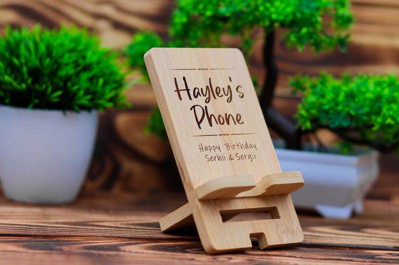 May include: A wooden phone stand with the text "Hayley's Phone" and "Happy Birthday Serhii & Sergii" engraved on it. The stand is made of natural bamboo and has a simple design.