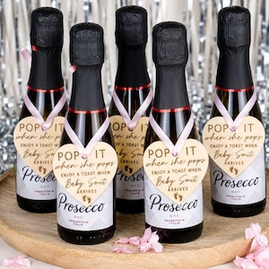 Può includere: Cinque bottiglie di Prosecco in miniatura con colli avvolti in carta stagnola nera, ciascuna dotata di un nastro rosa chiaro che sostiene un'etichetta in legno a forma di cuore. Le etichette riportano il testo "POP IT when she pops" e "ENJOY A TOAST WHEN Baby Smit ARRIVES."
