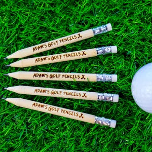 Op de afbeelding: Vijf houten potloden met de tekst "ADAM'S GOLF PENCILS X" erop gegraveerd. De potloden liggen op een groene grasmat naast een witte golfbal.
