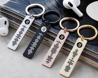 May include: Four rectangular metal keychains in silver, black, rose gold, and gold tones. Each keychain features a circular ring and a flat pendant engraved with a black Spotify logo and a unique sound wave pattern, arranged in a row.