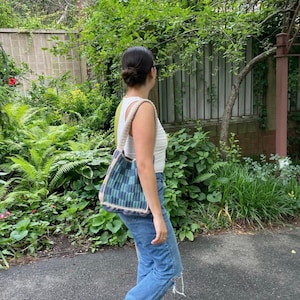 May include: A person wearing a white tank top and blue jeans is carrying a blue and green crocheted tote bag.