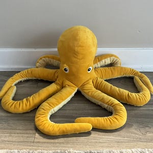 May include: A large, plush octopus toy in a vibrant yellow color. The stuffed animal has eight long, curved tentacles and a rounded head with two black and white eyes. The underside of the tentacles is a lighter cream color.