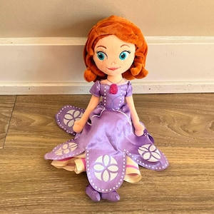 May include: A plush doll featuring auburn hair, bright blue eyes, and a soft fabric construction. The doll wears a purple satin dress with white floral-patterned petal overlays, a pink circular pendant at the neckline, and matching purple shoes.