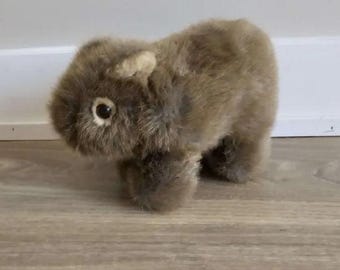 May include: A plush toy shaped like a wombat with thick, textured, light brown fur. The stuffed animal features a rounded body, short legs, small ears, and a single visible dark eye with a light-colored ring around it, captured in a side profile.