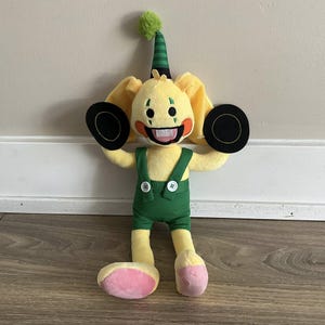 May include: A plush toy character with yellow fur, large floppy ears, and a green and black striped party hat. The character wears green dungarees and holds two black circular objects. The toy has a wide, toothy grin and pink accents on its feet.