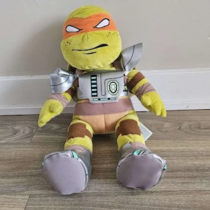 May include: A plush toy of a Teenage Mutant Ninja Turtle, Donatello, wearing a silver and green shell and a brown and yellow bandana. The toy has a green face with orange eyes and a serious expression.