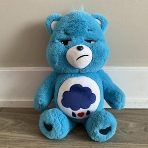 May include: A blue plush Care Bear with a downcast expression. The bear features a white belly with a dark blue cloud and red heart design. The bear is sitting on a wooden floor.